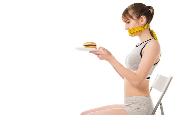 Mujer joven con la boca atada en cinta métrica sosteniendo hamburguesa en placa aislada en blanco - foto de stock