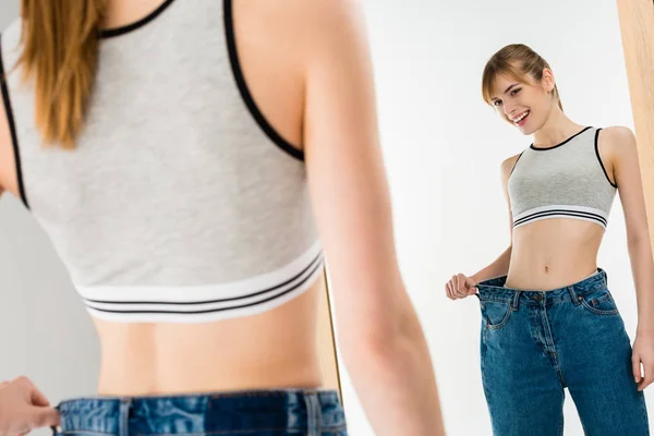 Mujer delgada feliz en pantalones vaqueros de gran tamaño mirando espejo aislado en blanco — Stock Photo
