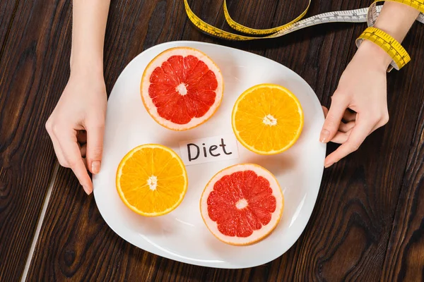 Cropped shot of woman with measuring tape on hand holding plate with halved citrus fruits and diet inscription — Stock Photo