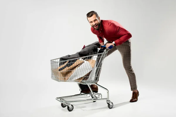 Uomo allegro con carrello pieno di shopping bag e giacca isolati su grigio — Foto stock