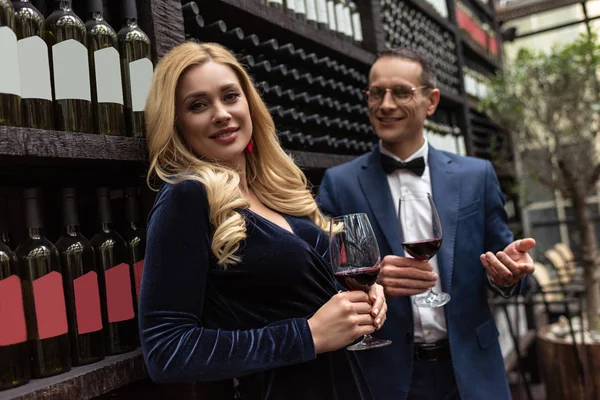 Feliz pareja adulta bebiendo vino delante de los estantes de almacenamiento de vino - foto de stock