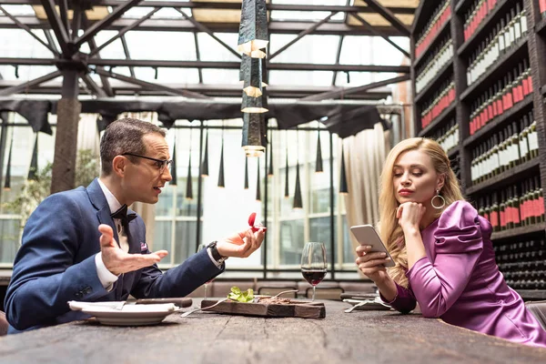 Hombre proponiendo a su novia mientras ella usa el teléfono inteligente con expresión aburrida en el restaurante - foto de stock