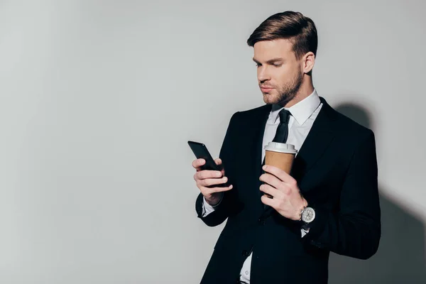 Stylish thoughtful businessman in suit holding paper cup and looking at smartphone screen on white background — Stock Photo