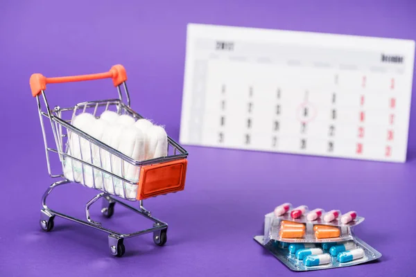 Tampons in small shopping cart, pills and calendar on purple — Stock Photo