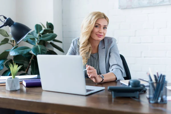 Feliz mujer de negocios sentada cerca de la computadora portátil y mirando a la cámara - foto de stock