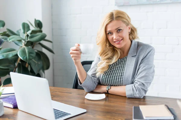 Happy businesswoman holding cup of coffee in office and looking at camera — Stock Photo