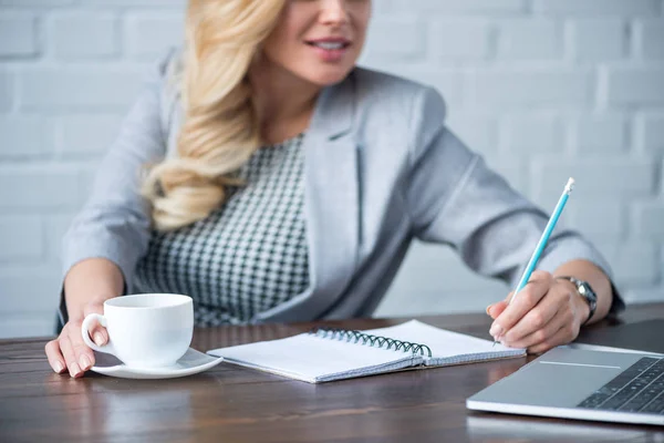 Abgeschnittenes Bild einer Geschäftsfrau, die etwas ins Notizbuch schreibt und eine Tasse Kaffee hält — Stockfoto