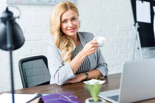 Sonriente mujer de negocios sosteniendo taza de café en la oficina y mirando a la cámara - foto de stock
