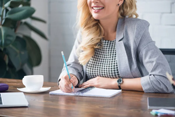 Imagem recortada de mulher de negócios sorridente escrevendo algo para notebook com lápis no escritório — Fotografia de Stock