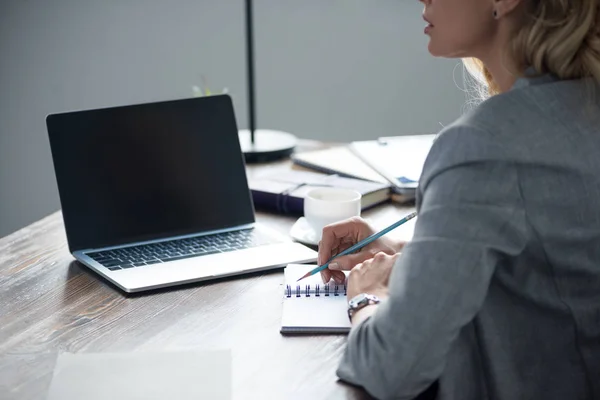 Image recadrée de femme d'affaires écrit quelque chose pour ordinateur portable avec crayon dans le bureau — Photo de stock