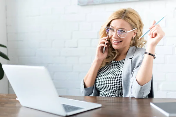 Счастливая деловая женщина разговаривает по смартфону в офисе с ноутбуком — стоковое фото