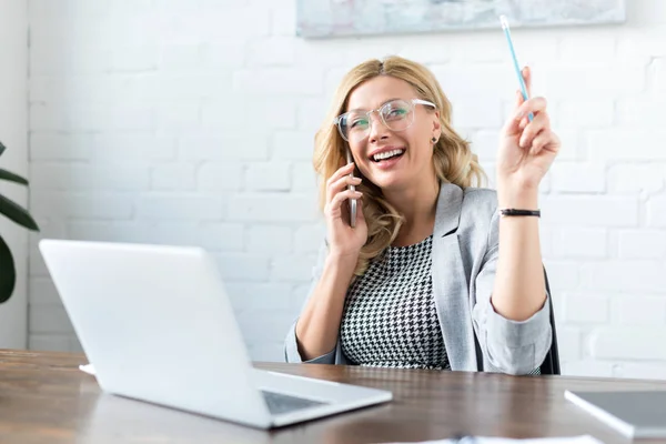 Lächelnde Geschäftsfrau, die im Büro per Smartphone und Laptop spricht — Stockfoto