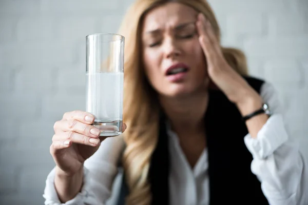 Mujer de negocios con dolor de cabeza y sosteniendo un vaso de agua con medicamentos — Stock Photo
