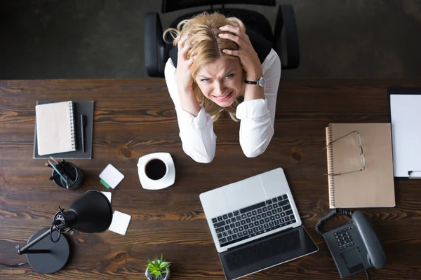 Blick von oben auf gestresste Geschäftsfrau, die den Kopf mit den Händen berührt und im Büro in die Kamera schaut — Stockfoto