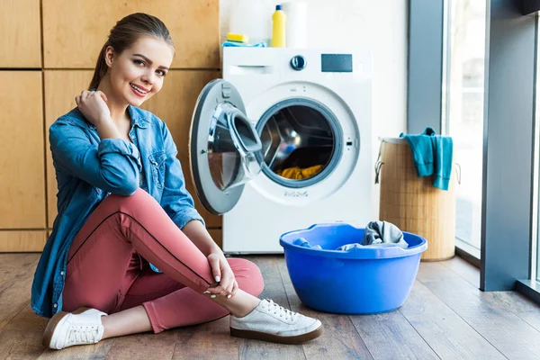 Bela jovem mulher sorrindo para a câmera enquanto sentado perto da máquina de lavar roupa e bacia de plástico com lavanderia — Fotografia de Stock