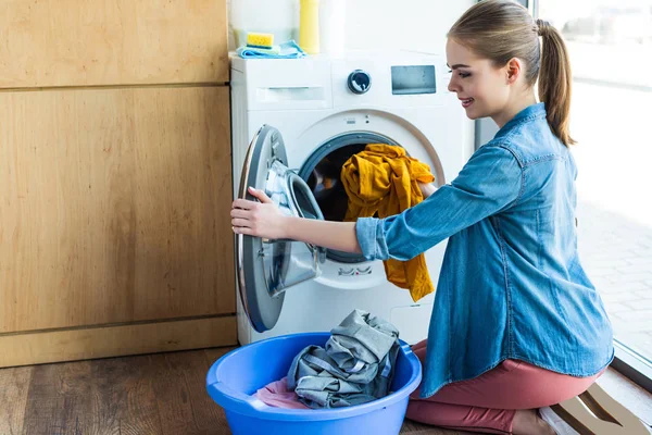 Jeune femme souriante prenant la lessive de la machine à laver dans un bassin en plastique — Photo de stock