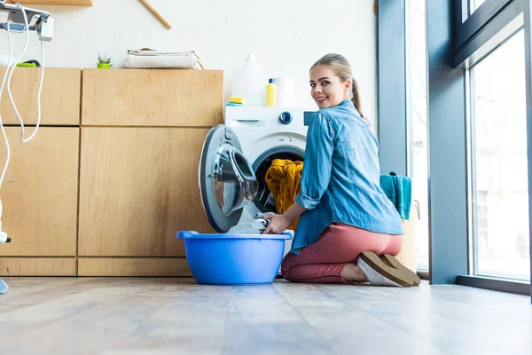 Jeune femme prenant la lessive de la machine à laver et souriant à la caméra — Photo de stock