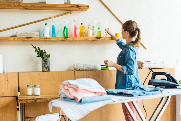 Young woman choosing cleaning fluid while ironing clothes at home — Stock Photo