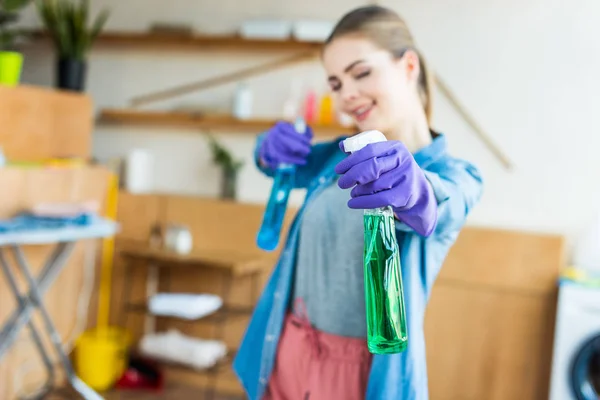 Nahaufnahme einer lächelnden jungen Frau in Gummihandschuhen, die Sprühflaschen mit Reinigungsflüssigkeit in der Hand hält — Stock Photo