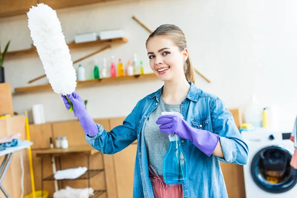 Sorrindo jovem mulher em luvas de borracha segurando spray garrafa e espanador — Fotografia de Stock