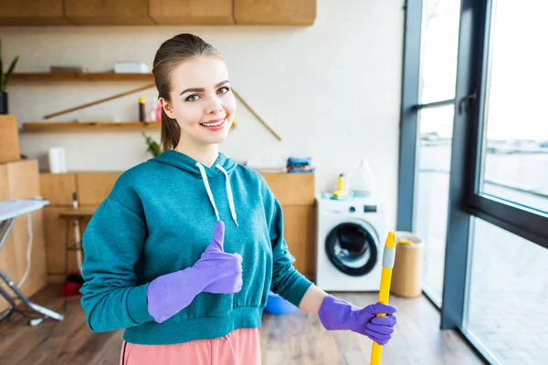 Sorrindo jovem mulher limpeza casa com esfregona e mostrando polegar para cima — Fotografia de Stock