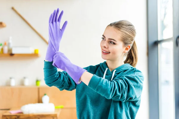 Belle jeune femme souriante portant des gants en caoutchouc lors du nettoyage de la maison — Photo de stock