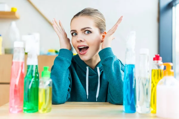 Sorprendió a la joven mujer mirando a la cámara mientras estaba sentada en la mesa con botellas de plástico con productos de limpieza - foto de stock