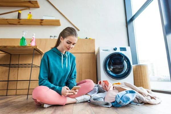 Улыбающаяся молодая женщина с помощью смартфона, сидя с одеждой возле стиральной машины — стоковое фото