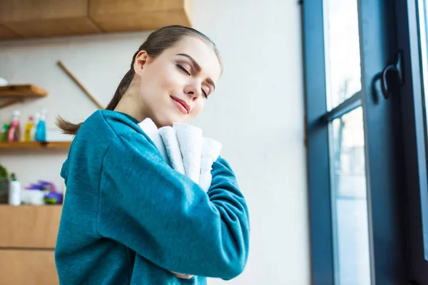 Lächelnde junge Frau mit geschlossenen Augen, die zu Hause saubere Handtücher umarmt — Stockfoto