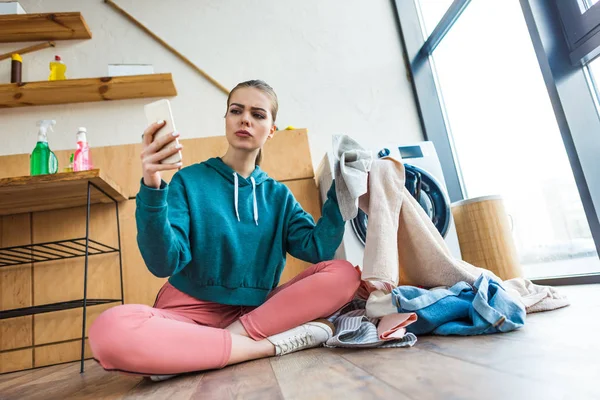 Young woman using smartphone while holding clothes and sitting near washing machine — Stock Photo