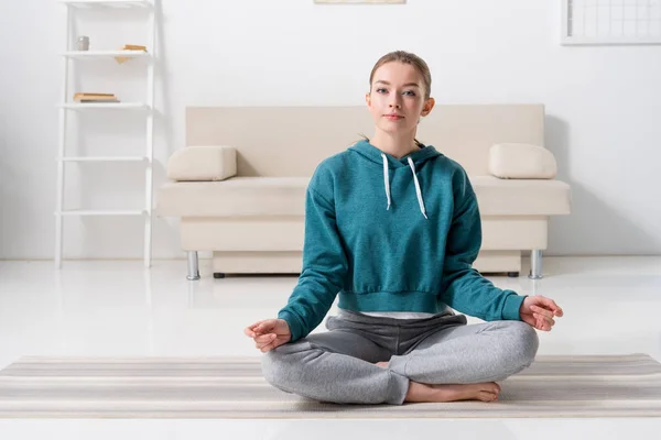Chica sentada en posición de loto en estera de yoga en casa y mirando a la cámara - foto de stock