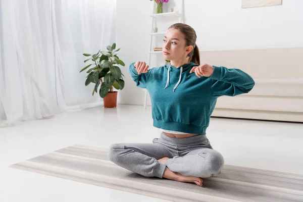 Chica sentada en posición de loto y estirándose en la esterilla de yoga en casa - foto de stock