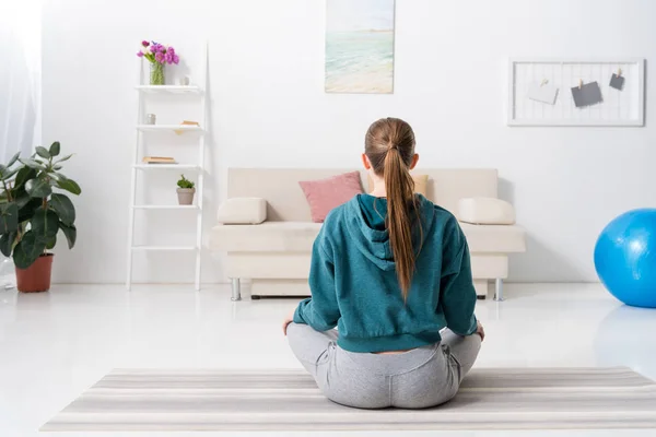 Vista trasera de la chica sentada en posición de loto en la esterilla de yoga en casa - foto de stock