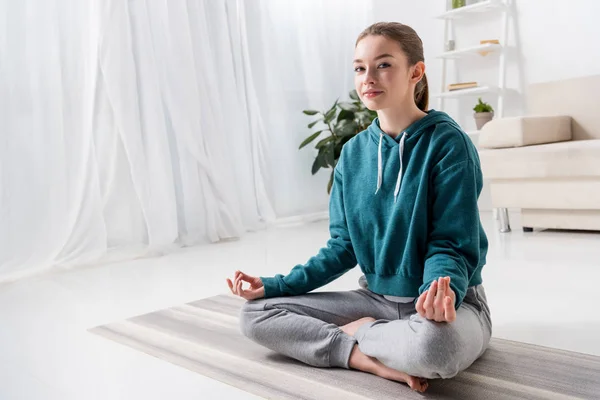 Hermosa chica sentada en posición de loto en estera de yoga y mirando a la cámara en casa - foto de stock