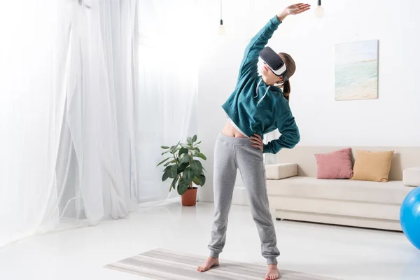 Menina de pé e alongamento com fone de ouvido realidade virtual no tapete de ioga em casa — Fotografia de Stock