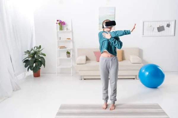 Девушка протягивает руки с гарнитурой виртуальной реальности на коврик для йоги дома — стоковое фото
