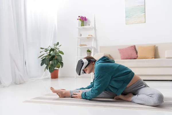 Ragazza atletica che si estende con auricolare realtà virtuale su tappetino yoga a casa — Foto stock