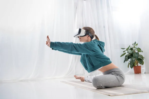 Vista lateral de chica estirándose con auriculares de realidad virtual en esterilla de yoga en casa - foto de stock