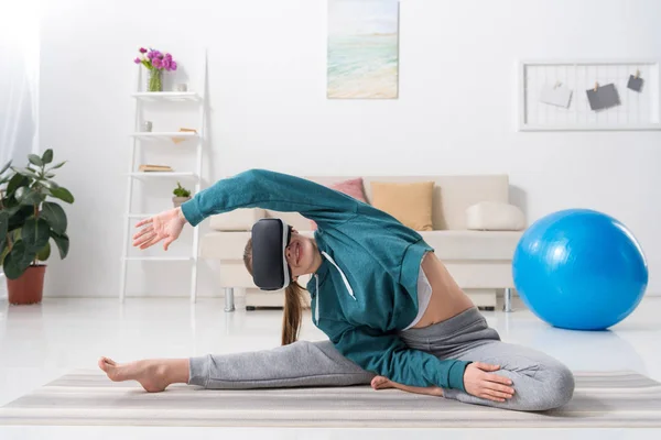 Девушка растягивая тело с гарнитурой виртуальной реальности на циновке yoga дома — стоковое фото
