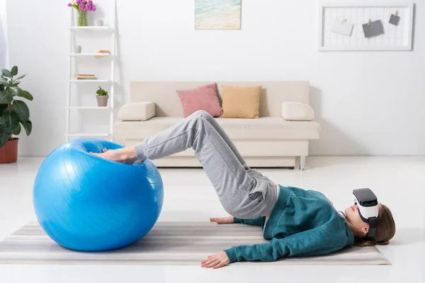 Vista lateral de la chica haciendo ejercicio con la pelota de fitness y en realidad virtual auriculares en casa - foto de stock