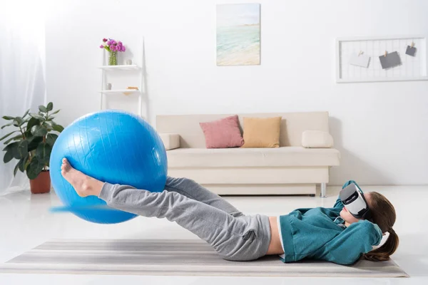Chica haciendo ejercicio con pelota de fitness y en realidad virtual auriculares en casa - foto de stock