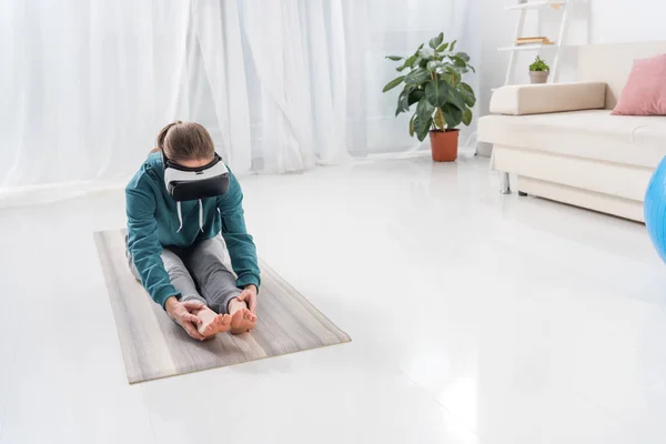 Девушка растягивается назад с гарнитурой виртуальной реальности на циновке йоги дома — стоковое фото