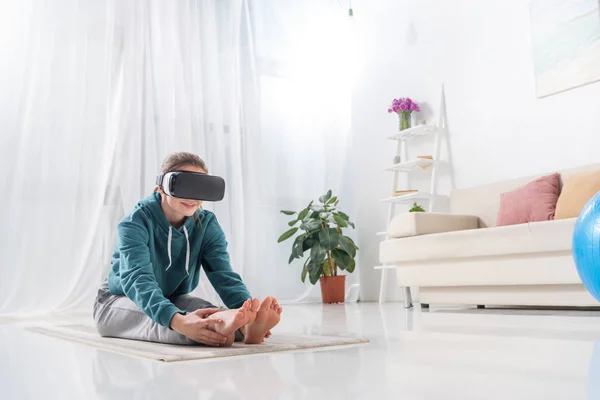 Menina alongamento com realidade virtual fone de ouvido no tapete de ioga em casa — Fotografia de Stock