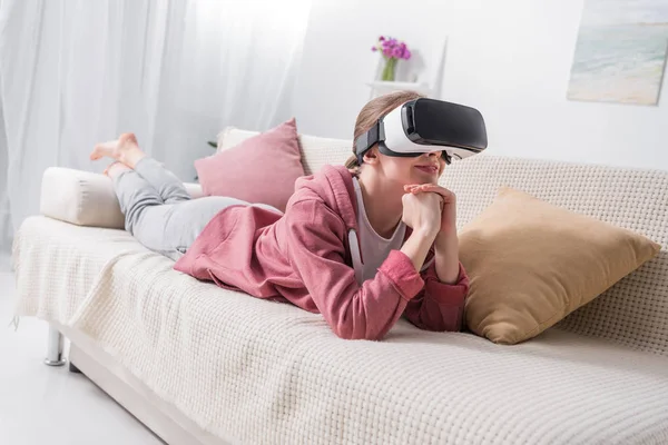Счастливая девочка лежит на диване и смотрит что-то с гарнитурой виртуальной реальности дома — стоковое фото