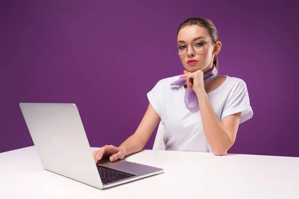 Pensive girl using laptop and looking at camera isolated on purple — Stock Photo