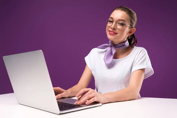 Усміхнена дівчина використовує ноутбук і дивиться на камеру ізольовано на фіолетовому — стокове фото