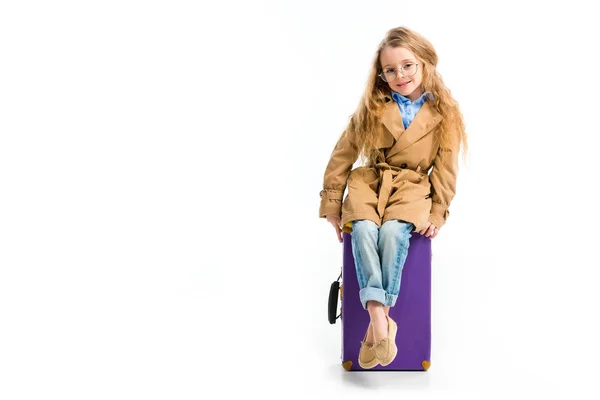 Enfant élégant en lunettes et trench coat assis sur une valise isolée sur du blanc — Photo de stock