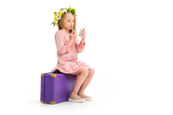 Vista laterale del bambino seduto sulla valigia viola e facendo il trucco dal rossetto isolato su bianco — Foto stock