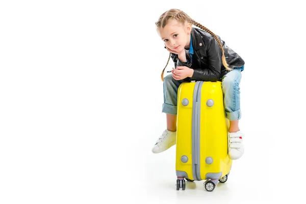 Petit enfant avec des nattes tenant la main sur le menton et assis sur la valise de roue isolé sur blanc — Photo de stock
