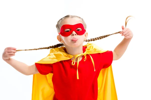 Pequeña supergirl en capa amarilla y máscara roja para los ojos mostrando la lengua y sosteniendo coletas aisladas en blanco - foto de stock
